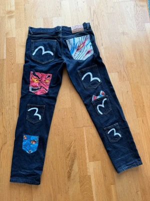 Mörkblå jeans med broderade masker - Unika mörkblå jeans från Evisu med broderade japanska masker och vita måsloggor på fickorna. Jeansen har raka ben och detaljer i rött, blått och vitt. Tillverkade i kraftig denim med läderpatch i midjan. Perfekta för dig som vill sticka ut med din stil.