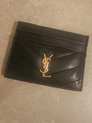 Svart kortfodral från Saint Laurent - Snyggt svart kortfodral från Saint Laurent i läder med guldfärgad YSL-logga framtill. Fodralet har flera kortfack och stilrena, diagonala sömmar som ger en lyxig känsla. Perfekt för dig som vill ha ett elegant och smidigt sätt att bära dina kort.