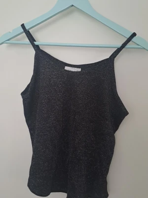 Glittrig svart linnetopp - Svart linnetopp från KIT med tunna axelband och glittrigt material. Materialet är 'lent' på framsidan, dvs är inte strävt som många andra plagg med glitter. Barnstorlek ska passa xs-s, eventuellt m. 🩷