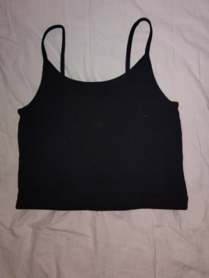 Svart ribbad linnetopp - En enkel och stilren svart linnetopp med smala axelband och ribbad struktur. Toppen har en croppad passform och är perfekt att matcha med höga jeans eller kjol. Materialet känns mjukt och stretchigt, vilket gör den bekväm att bära hela dagen.