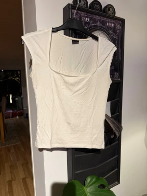 Vit topp med fyrkantig ringning - Stilren vit topp med breda axelband och fyrkantig ringning. Tunn och mjuk i materialet, perfekt för lager på lager eller sommaroutfits. Enkel och tidlös design som passar till det mesta. Den är lite noppring men inget som märks jätte mycket 