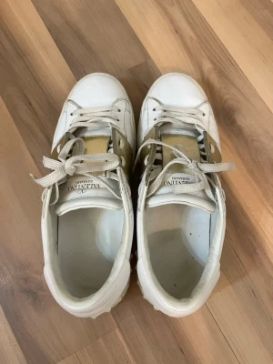 Valentino vita sneakers med gulddetalj - Snygga vita sneakers från Valentino med bred guldfärgad rem över sidan och klassiska nitar på sulan. Skorna har vita skosnören och är tillverkade i läder. Perfekta för dig som vill ha en exklusiv touch på din outfit.