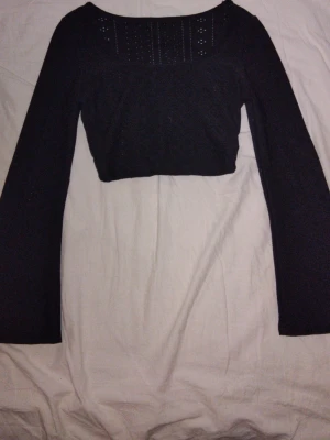 Topp - Svart croppad långärmad topp i storlek S. Toppen har fyrkantig ringning och långa utsvängda ärmar med fina detaljer som ger ett elegant utseende. Även baktill finns dekorativa detaljer som gör toppen extra speciell. Stretchigt material, kroppsnära passform och i bra skick.