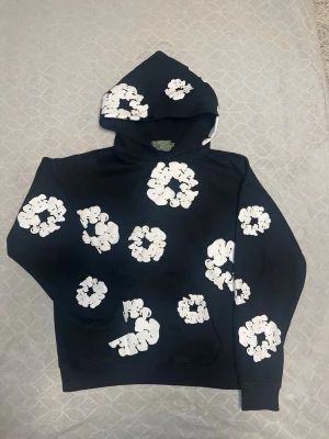 Svart hoodie med vita blommönster - Cool svart hoodie med stora vita blommönster i grafisk stil över hela plagget, även på huvan. Hoodien har klassisk passform, känguruficka framtill och ribbade muddar. Perfekt för dig som vill sticka ut med en unik och trendig look.