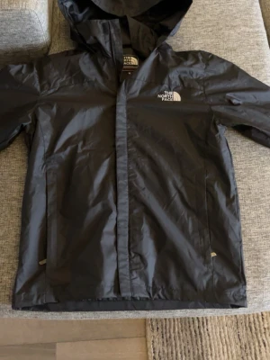 Svart regnjacka från The North Face - Snygg svart regnjacka från The North Face med huva och dragkedja framtill. Jackan har två sidofickor och logga på bröstet samt ryggen. Tillverkad i ett vattenavvisande material som passar perfekt för regniga dagar.