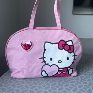 Hello Kitty väska - Söt rosa handväska med Hello Kitty-tryck på framsidan, dekorerad med små vita hjärtan och quiltad yta. Väskan har två handtag, dragkedja och ett hjärtdetalj med rosett. Insidan är röd med samma hjärtmönster. Perfekt för dig som gillar kawaii och vill sticka ut. Men den är även sliten på sidorna på väskan som du kan se på bilderna 