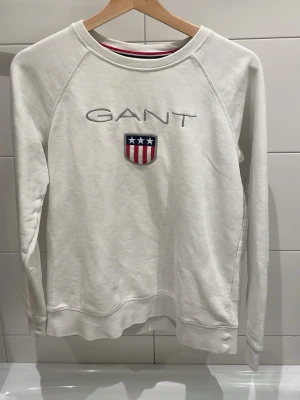 Vit sweatshirt från GANT - Vit sweatshirt från GANT med broderad logotyp och emblem med stjärnor och ränder på bröstet. Tröjan har rund halsringning, långa ärmar och ribbade muddar. Materialet är mjukt och bekvämt, perfekt för en avslappnad stil. Har dock 2 fläckar som jag tror går att tvätta bort 