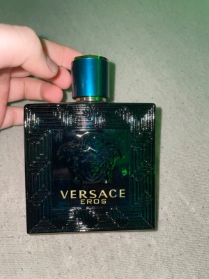 Versace Eros parfym flaska - Snygg parfymflaska från Versace, modellen Eros. Flaskan är mörkgrön med gulddetaljer och har ett coolt Medusa-huvud på både framsidan och korken. Fyrkantig form i glas med en metallisk blågrön kork. Perfekt för dig som gillar exklusiva dofter. UNGEFÄR 96-97 ML KVAR