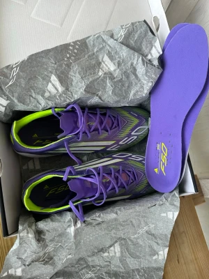 Adidas F50 lila och gröna fotbollsskor - Säljer ett par Adidas F50 fotbollsskor i en riktigt snygg lila och grön färgkombination med vita detaljer. Skorna har en modern, strömlinjeformad design och är tillverkade i syntetmaterial för lätt vikt och bra bollkontroll. Perfekta för dig som vill sticka ut på planen. Använde en gång då de skulle testas, men märkte att de inte passade min fot.