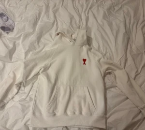 Vit hoodie med rött hjärta - Säljer en stilren vit hoodie från AMI Paris med deras ikoniska röda hjärta broderat på bröstet. Hoodien har huva med dragsko, magficka och ribbade muddar. Perfekt för dig som gillar enkel men snygg streetwear. Hoodien skulle jag påstå är storleken M/L något där emellan. Kan gå ned i pris vid snabb affär.