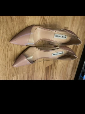 Nude pumps med transparent detalj Steve Madden - Säljer ett par snygga pumps från Steve Madden i nude färg med spetsig tå och transparenta sidor i plast. Skorna har en elegant och modern look med lackad finish framtill och klassisk klack. Perfekta för dig som vill sticka ut med stilrena detaljer.