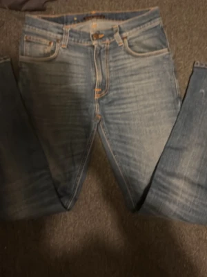 Blå jeans från Nudie Jeans - Säljer ett par klassiska blå jeans från Nudie Jeans med snygg slitning och kontrastsömmar. Modellen har fem fickor, gylf med knapp och dragkedja samt Nudie-logga på bakfickan. Perfekta till en avslappnad stil.