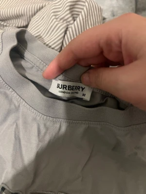 Grå Burberry t-shirt med broderi - Säljer en grå t-shirt från Burberry med stort broderat Burberry London England-tryck på bröstet. T-shirten har klassisk passform och rund hals. Broderiet har inslag av Burberrys ikoniska rutmönster i bokstäverna. Perfekt för dig som gillar stilrena märkesplagg.