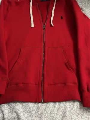 Polo Ralph Lauren hoodie  - Röd hoodie med dragkedja från Polo Ralph Lauren. Klassisk design med vit dragsko i huvan, två stora fickor fram och broderad marinblå logga på bröstet. Tillverkad i mjuk bomull för skön känsla och avslappnad stil.