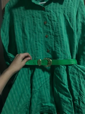 Grön kortklänning med bälte - Snygg grön kortklänning med strukturmönster, träknappar framtill och matchande grönt bälte med gulddetaljer. Klänningen har lång ärm och en avslappnad passform som ger en cool och trendig look. Jätte fin för sommaren använt 1 gång  köptes utomlands kommer inte ihåg vart i från ..😊