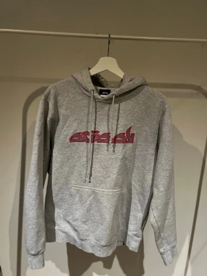 Grå hoodie från Stüssy med tryck - Säljer en grå hoodie från Stüssy i storlek S. Hoodien har ett fett rött tryck på bröstet, klassisk känguruficka och justerbar huva med snören. Tillverkad i mjukt bomullsmaterial som är skönt att ha på sig.