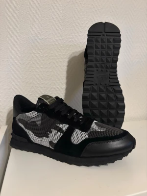 Valentino svart/vit camo sneakers - Snygga sneakers från Valentino med svart och vit camo-mönster. Skorna har mesh och mocka detaljer, samt en robust svart gummisula med mönstrad undersida. Klassisk låg modell med svarta skosnören och logotyp på plösen.