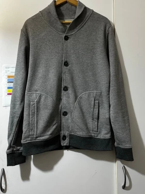 Grå cardigan  - Snygg grå cardigan från Selected Homme med knäppning framtill och två stora fickor. Tröjan har ribbade muddar vid ärmslut och nederkant samt en hög krage. Perfekt för lager på lager och enkel att matcha med olika outfits.
