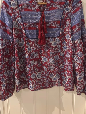 Blommig boho-blus från Indiska - Blus i boho-stil från Indiska med röd botten och blåvita blommor. Har långa ärmar, dekorativa blå ränder över axlar och bröst samt knytband med tofsar vid halsen. Tillverkad i ett mjukt, vävt material som känns luftigt och skönt.