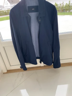Mörkblå vindjacka från BOSS - Snygg mörkblå vindjacka från BOSS med dragkedja, krage och två sidofickor. Jackan har minimalistisk design och justerbar nederkant med dragsko. Perfekt för dig som vill ha en stilren och lätt jacka till vardags. Materialet känns tunt och smidigt.