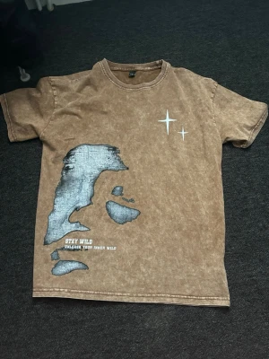 Brun t-shirt med tryck och text - Cool brun t-shirt med tvättad look och unikt tryck i blått och vitt framtill. Texten 'STAY WILD UNLEASH YOUR INNER WILD' syns nedtill. Två vita stjärnor på bröstet ger extra detalj. Perfekt för dig som gillar streetwear vibes.