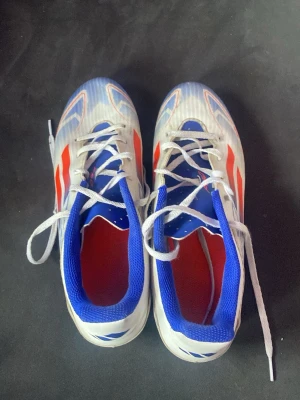 Adidas blåvita fotbollsskor - Säljer ett par Adidas fotbollsskor i vitt, blått och rött. Skorna har klassisk snörning, låg profil och en ovandel i syntetmaterial. Insidan är blå och röd, och yttersulan är designad för bra grepp på planen. Perfekta för dig som vill sticka ut på matchen!
