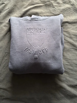 Grå Malmö FF hoodie  - Säljer en grå hoodie från Malmö FF med broderad logga och text på bröstet. Hoodien har en klassisk känguruficka framtill, ribbade muddar och en skön huva. Perfekt för dig som vill visa ditt stöd för laget på ett stilrent sätt.