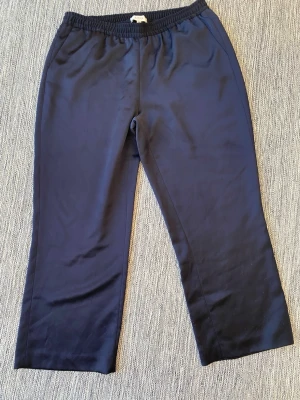 Mörkblå kostymbyxor från Gigue - Mörkblå kostymbyxor från Gigue med rak passform och elastisk midja. Byxorna har en stilren look och är tillverkade i ett mjukt, lätt tyg som ger en clean vibe. Perfekta för dig som gillar enkel och snygg stil. Eleganta, mörkblå glansiga byxor med resår i midjan. Byxorna har en avslappnad passform och är perfekta för en bekväm men stilfull look.   Sparsamt använda och i fint begagnat skick   Mått Midjan: ca 43 cm Längd fr grenen o ner: ca 68 cm  100% polyester   Kommer fr ett djur/rökfritt hem