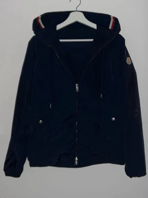 Mörkblå windbraker från Moncler  - Snygg marinblå vindjacka från Moncler med huva och dragkedja framtill. Jackan har två fickor med tryckknappar, dragsko i huvan och Moncler-logga på ärmen. Röd och vit randig detalj på huvan ger en sportig touch. Såklart äkta. Orginal påse tillkommer. OBS kvitto tillkommer inte. Priset är såklart diskuterbart 