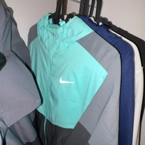 Turkos Nike vindjacka med huva - Snygg turkos och grå vindjacka från Nike med vit logga på bröstet. Jackan har huva med dragsko och dragkedja framtill. Perfekt för dig som gillar sportig stil och vill sticka ut med färg.