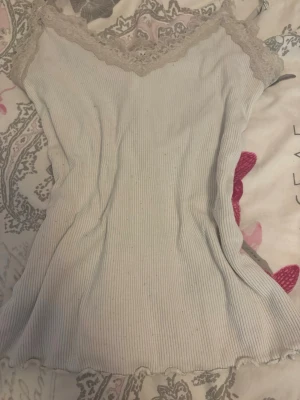 Vit ribbad linne med spets - Säljer ett vit ribbat linne med tunna axelband och vacker beige spetsdetalj vid urringningen. Linnet har en mjuk och stretchig känsla, perfekt för avslappnade dagar hemma. Materialet känns lätt och luftigt, och nederkanten har en vågig finish.