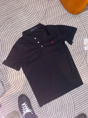 Svart pikétröja från Polo Ralph Lauren - Klassisk svart pikétröja från Polo Ralph Lauren med korta ärmar och tre vita knappar framtill. Tröjan har den ikoniska röda logotypen broderad på bröstet och är tillverkad i mjuk bomull. Perfekt för en clean och stilren look. Helt ny oanvänd!