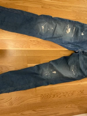 Blå slitna jeans från Replay - Säljer ett par blå jeans från Replay med slitningar och distressed detaljer på både fram- och baksida. Klassisk femficksmodell med raka ben och normal midja. Jeansen är i denim och har snygga slitna partier för en trendig look.