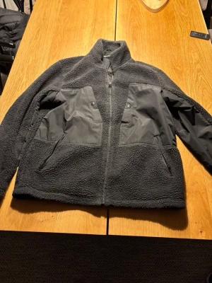 Mörkgrå teddyjacka med detaljer - Säljer en mörkgrå teddyjacka med dragkedja framtill och höga fickor med lock och dragkedja. Jackan har kontrasterande tygdetaljer på bröstet och ena ärmen, samt hög krage för extra värme. Perfekt för dig som gillar en stilren och mysig look.