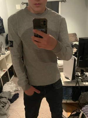 Grå långärmad tröja från Calvin klein  - Snygg grå långärmad tröja från Calvin Klein med klassisk patch på vänster ärm. Tröjan har rund halsringning och är tillverkad i ett mjukt, ribbat material som ger en clean och stilren look. Perfekt till jeans eller chinos för en avslappnad stil. Helt ny oanvänd 