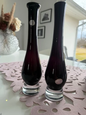 Orrefors glasvaser mörklila - Två eleganta glasvaser från Orrefors, designade av Nils Landberg. Vaserna är mörklila med klara glasfötter och har en smal, hög form som passar perfekt för enstaka blommor. Klassisk svensk design som blir ett snyggt inslag i hemmet.