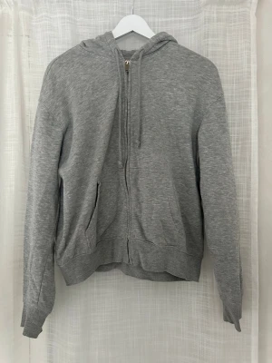 Grå zip hoodie från H&M - Klassisk grå zip hoodie från H&M med huva och dragsko. Tillverkad i mjuk bomullsblandning och har två fickor framtill samt ribbade muddar vid ärmar och nederkant. Perfekt för en avslappnad och enkel stil.