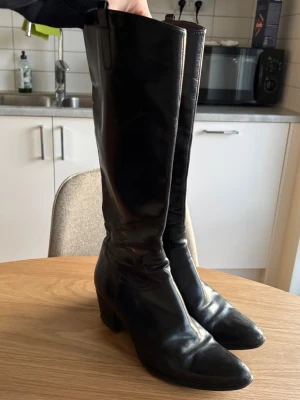 Svarta knähöga läderstövlar - Säljer ett par klassiska svarta knähöga stövlar i äkta läder med rundad tå och bred klack. Stövlarna har dragflikar upptill för enkel påtagning och en stilren, tidlös design som passar till många outfits.