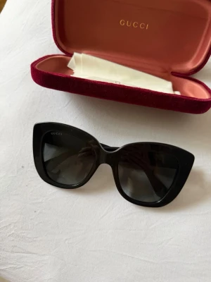 Gucci svarta cat eye solglasögon - Snygga svarta cat eye-solglasögon från Gucci med breda bågar och guldfärgad logga på sidorna. Kommer med originalfodral i röd sammet och putsduk. Jättefint skick!
