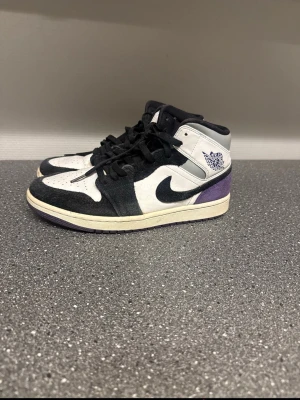 Nike Air Jordan 1 Mid svart/vit - Snygga Nike Air Jordan 1 Mid sneakers i svart, vitt och lila. Klassisk siluett med svart swoosh, vita paneler och lila detaljer vid hälen. Tillverkade i läder och mocka med snörning framtill och ikonisk Air Jordan-logga på sidan.