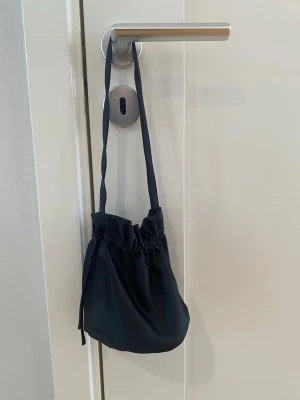 Svart satinpåse handväska - En svart handväska i mjukt satinliknande material med dragsko upptill och smal axelrem. Väskan har en rundad botten och enkel, stilren design som passar till många olika looks. Perfekt för dig som gillar minimalistiska accessoarer. Aftonväska svart glansigt tyg perfekt till balklänning eller aftonklänning.  Väskan är endast använd vid ett tillfälle och är i nyskick   Kommer från ett djur och rökfritt hem.