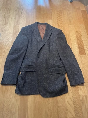 Mörkbrun kavaj från Hugo Boss - Stilren mörkbrun tweedkavaj från Hugo Boss med klassisk passform och två framfickor med lock. Kavajen har ett diskret fiskbensmönster och är helfodrad med brunt innerfoder. Perfekt för en dressad look och enkel att matcha. Storlek 46. Priset är inte hugget i sten!