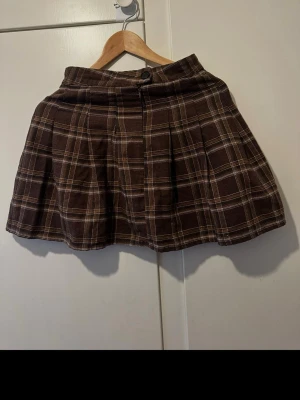 Brun rutig kortkjol med knapp - Kortkjol i brunt rutigt tyg med beige och vita ränder. Kjolen har en knapp framtill och lätt utställd passform. Perfekt för dig som gillar klassisk stil med en twist.