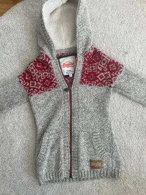 Grå stickad Superdry hoodie  - Stickad hoodie från Superdry i gråmelerat med rödvit nordiskt mönster över axlarna. Dragkedja framtill, två fickor och mjukt vitt foder i huvan. Perfekt för kalla dagar och riktigt snygg streetstil.