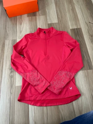  långärmad träningströja - Snygg rödrosa långärmad träningströja med halv dragkedja framtill och hög krage. Ärmarna har mönstrade detaljer i vitt och en liten logga vid nederkanten. Perfekt för löpning eller gym. Tillverkad i ett stretchigt funktionsmaterial.