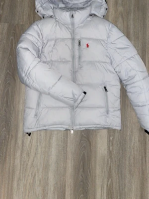 Ljusgrå dunjacka från Polo Ralph Lauren - Snygg ljusgrå dunjacka från Polo Ralph Lauren med huva och dragkedja framtill. Jackan har två sidofickor med dragkedja och den klassiska röda loggan broderad på bröstet. Perfekt för kalla vinterdagar och riktigt skön att bära. Bränn skada!!!!