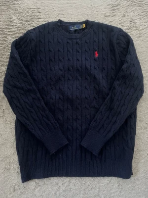 Mörkblå kabelstickad tröja Polo Ralph Lauren - Kabelstickad mörkblå tröja från Polo Ralph Lauren med rund halsringning och den klassiska röda logotypen broderad på bröstet. Tröjan har ribbade muddar vid ärmslut och nederkant. Perfekt för dig som gillar tidlös och stilren design.