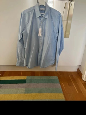 Ljusblå skjorta från John Henric - Klassisk ljusblå skjorta från John Henric med knappar framtill och lång ärm. Skjortan har en stilren krage och är tillverkad i bomull för en bekväm känsla. Perfekt för dig som gillar en clean och enkel look.