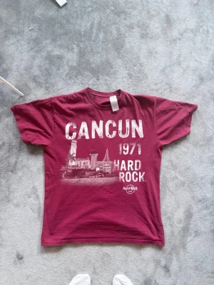 Vinröd Hard Rock Cancun t-shirt - Snygg vinröd t-shirt från Hard Rock Café med tryck av Cancun, 1971 och stadsmotiv på framsidan. Klassisk rund hals och avslappnad passform. Perfekt för dig som gillar ikoniska tryck och vill sticka ut lite extra. Passar bra till fester med rock tema🎸🎸