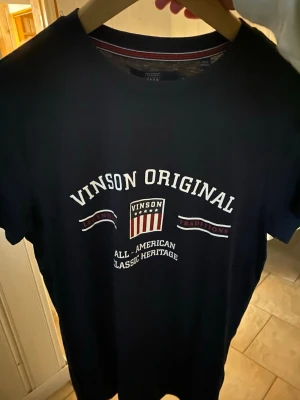 Svart t-shirt från Vinson Original - Snygg svart t-shirt från Vinson Original med tryck framtill i vitt och rött. Klassisk rund halsringning och korta ärmar. Perfekt för en avslappnad och stilren look.